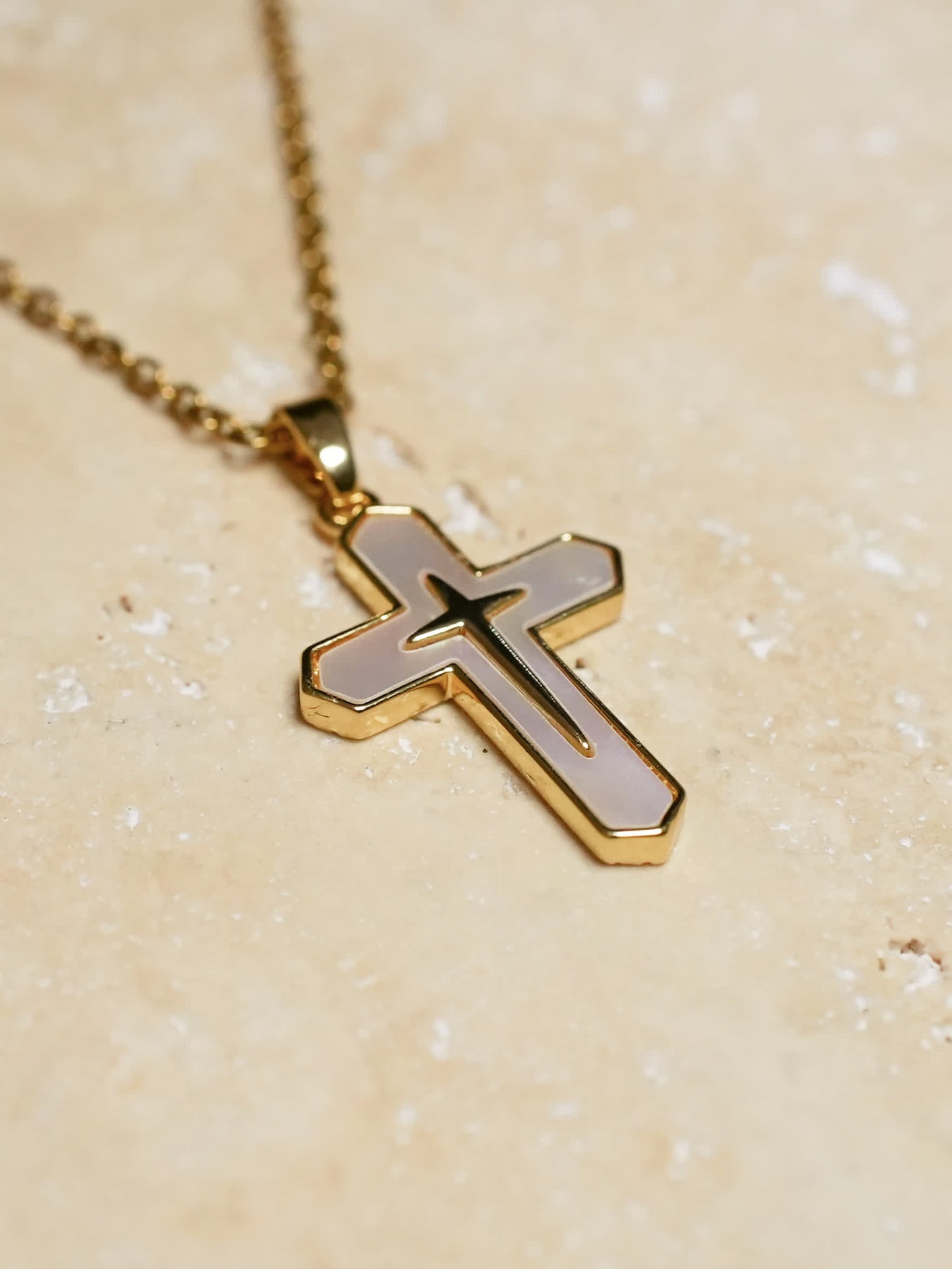 Gold and white cross necklace resting on a textured beige stone surface