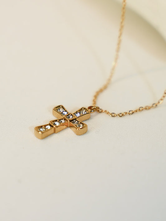 Gold cross necklace with inset zircons laid flat on a cream fabric surface, showing sparkle and clarity