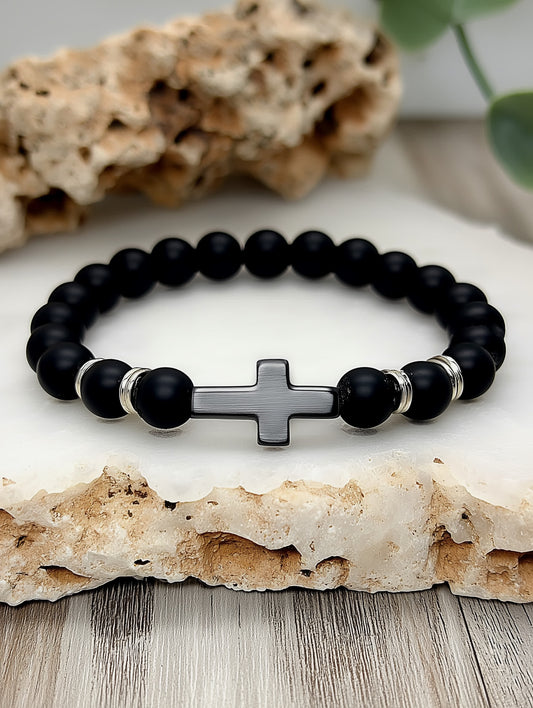 Angled lifestyle shot of the Covenant Shield Men's Cross Bracelet resting on textured white stone with natural rock in the background, showcasing a brushed stainless steel cross and polished gemstone beads.