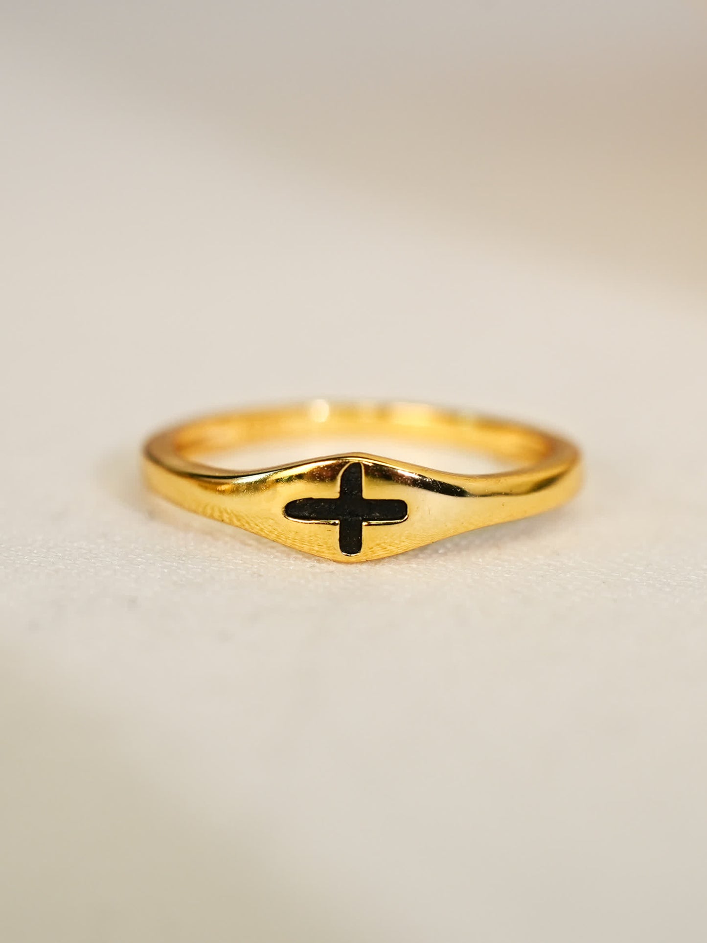 Sculpted signet-style gold ring with recessed black cross, displayed on soft ivory fabric