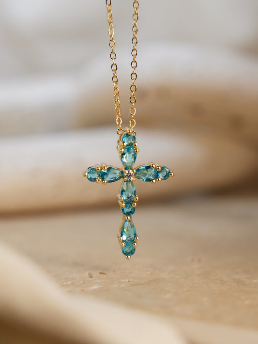 Blue cross necklace resting on travertine stone, highlighting teardrop gemstone sparkle and gold chain