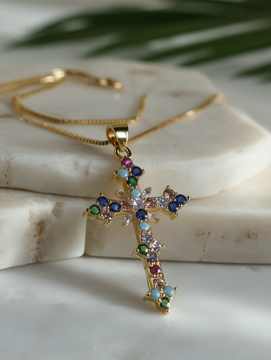 Close-up lifestyle photo of the rainbow cross pendant resting on marble with palm leaf in background, showing vibrant gemstone details