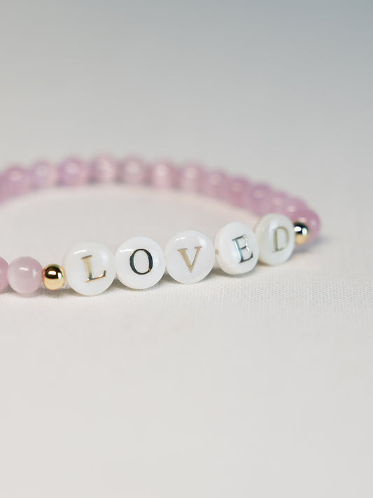 Close-up angled shot of the Heaven’s Whisper Faith Bracelet on a light background, highlighting the “LOVED” letters and soft pink beads.