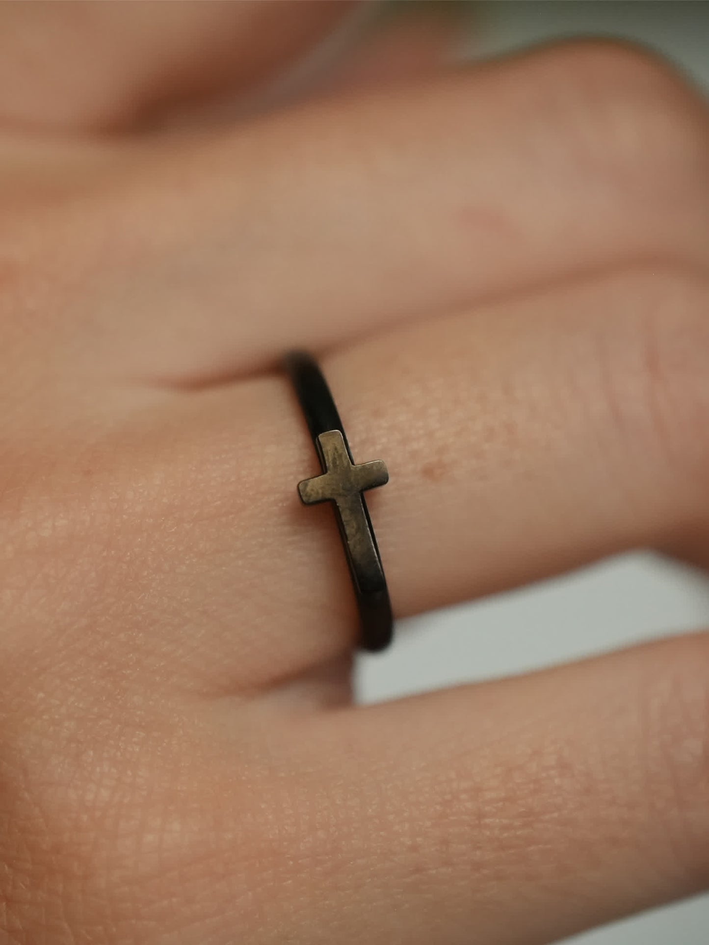 Macro side view of the woman’s cross ring, highlighting the antique bronze-tone cross detail against the matte black band on the finger.