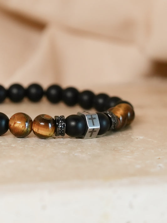 Close-up angled view of the Lion's Peace Men's Religious Bracelet lying flat on a beige stone surface, showcasing the cross charm and polished tiger’s eye beads.