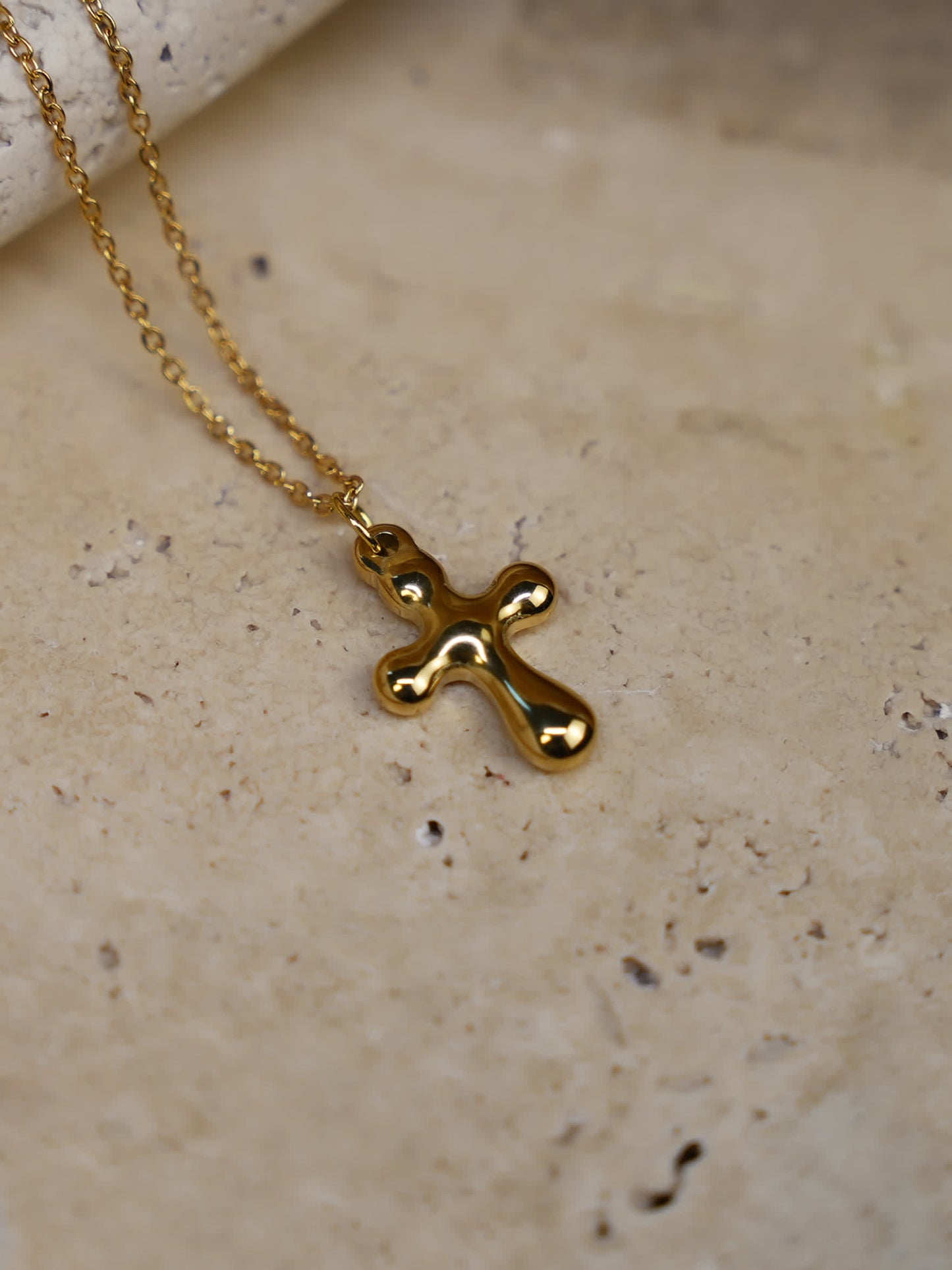 Flat-lay of the sculptural gold cross necklace on stone surface, showcasing the fluid abstract shape of the pendant