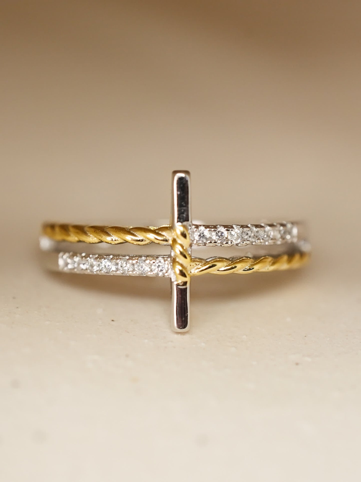 Stylized product shot of the Sacred Bind Ring on neutral stone surface, showing rhodium-plated band with cubic zircon details and gold accent wrap