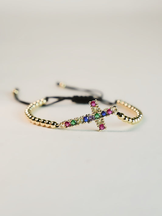 Angled studio shot of the Joy in Light Cross Bracelet on a clean off-white background, highlighting the sparkle of the colorful zircon stones set in a sideways gold cross.