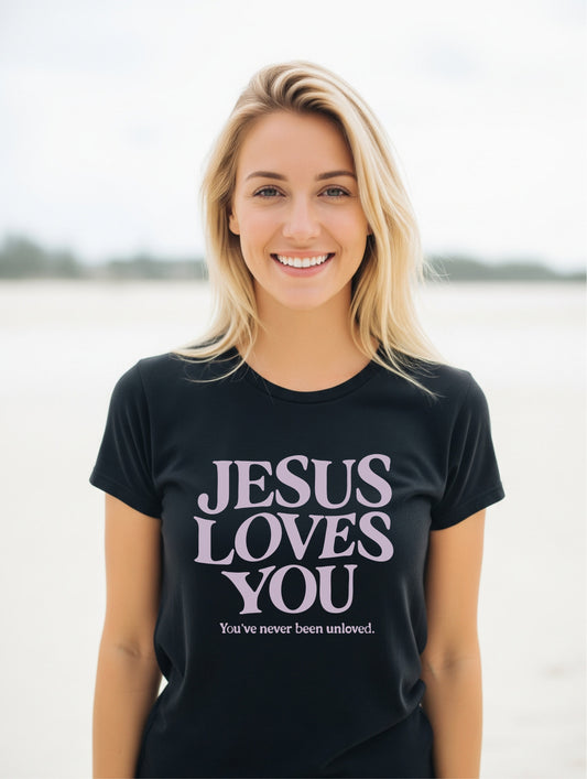 Woman wearing a black t-shirt with 'Jesus Loves You' text on a beach.