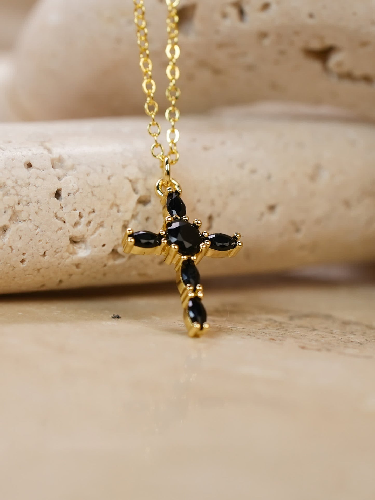 Obsidian marquise-cut gold cross pendant resting on beige travertine stone