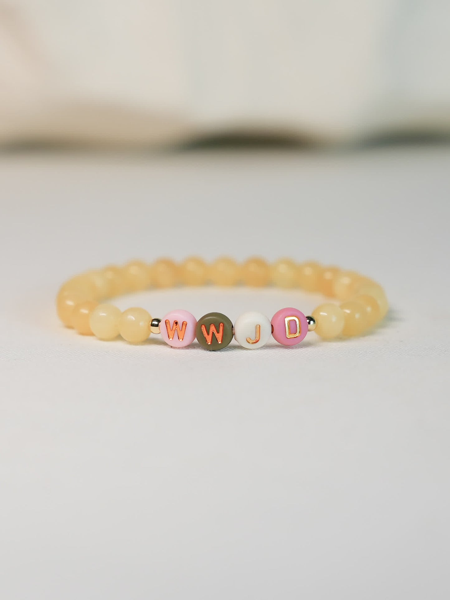 Front-facing image of the WWJD Bracelet on a soft white surface, showing warm yellow gemstone beads and multicolored WWJD letter beads with gold accents.