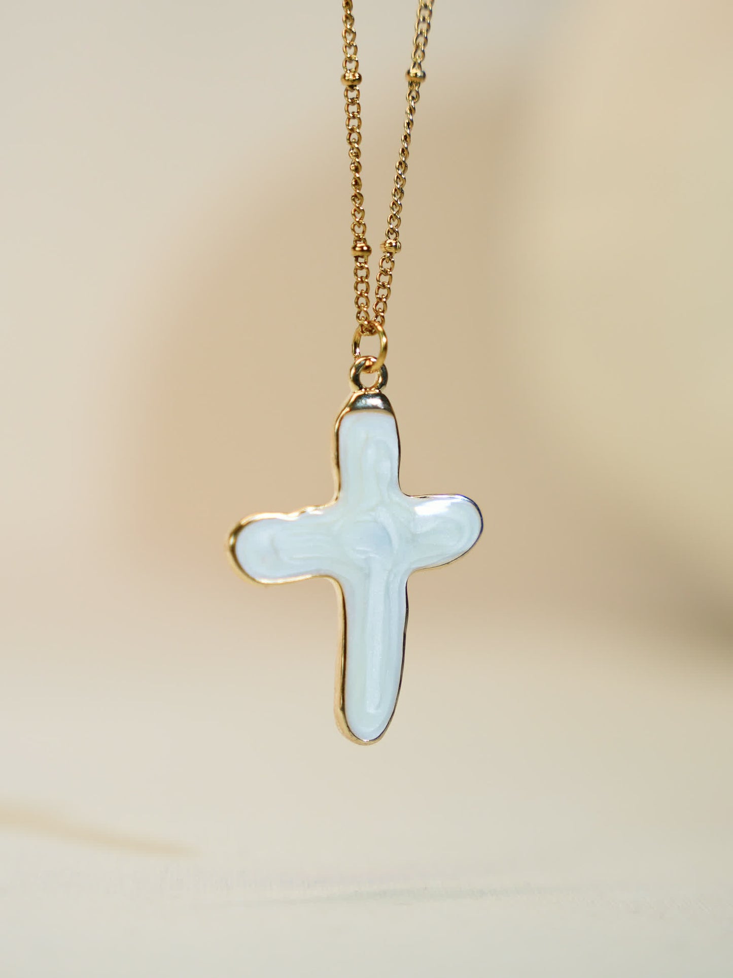 Studio shot of white shell cross necklace with polished gold chain – modern Christian accessory in minimal beige backdrop.