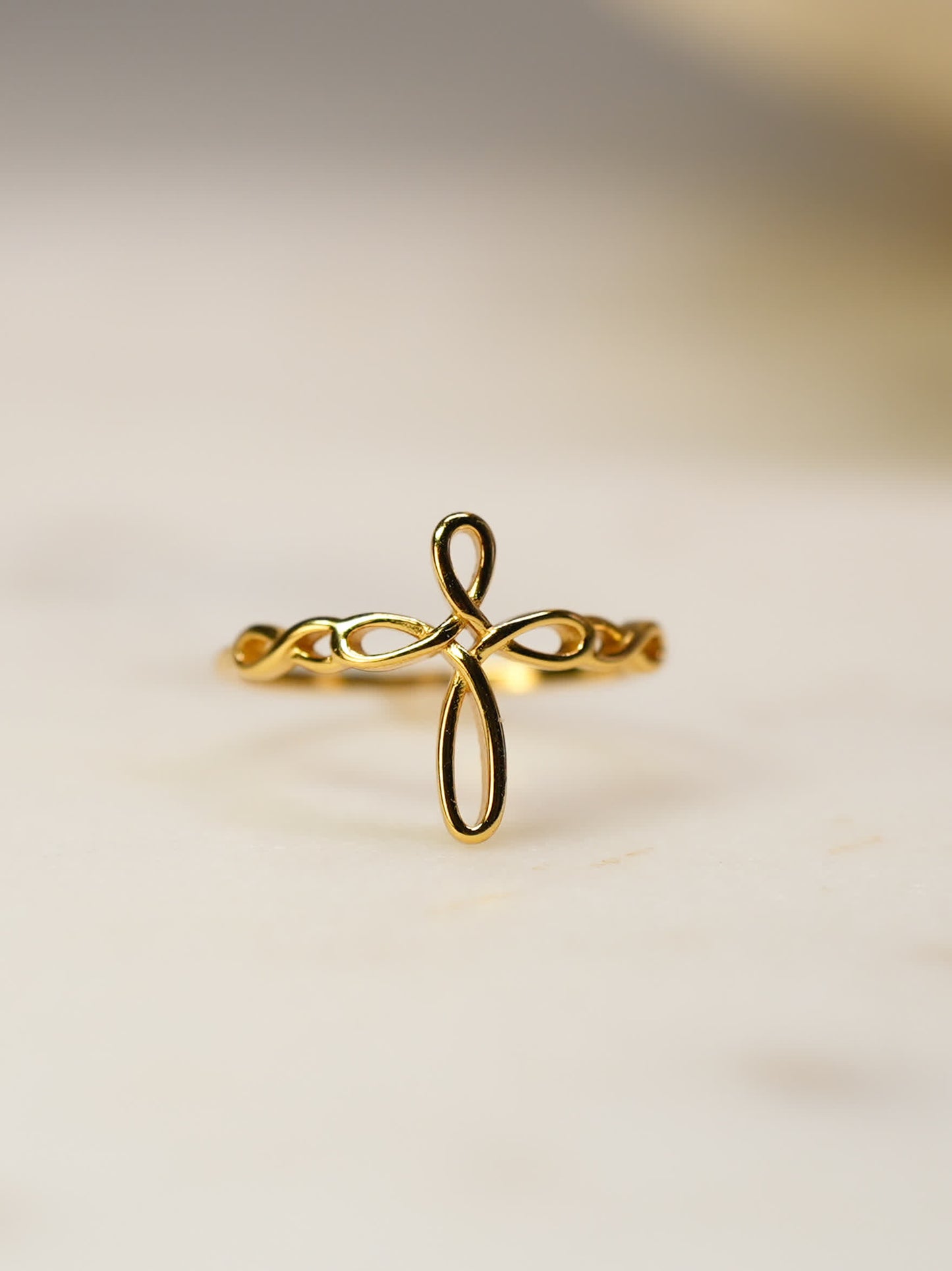 Close-up of gold ribbon faith ring resting on soft marble surface, highlighting flowing cross silhouette