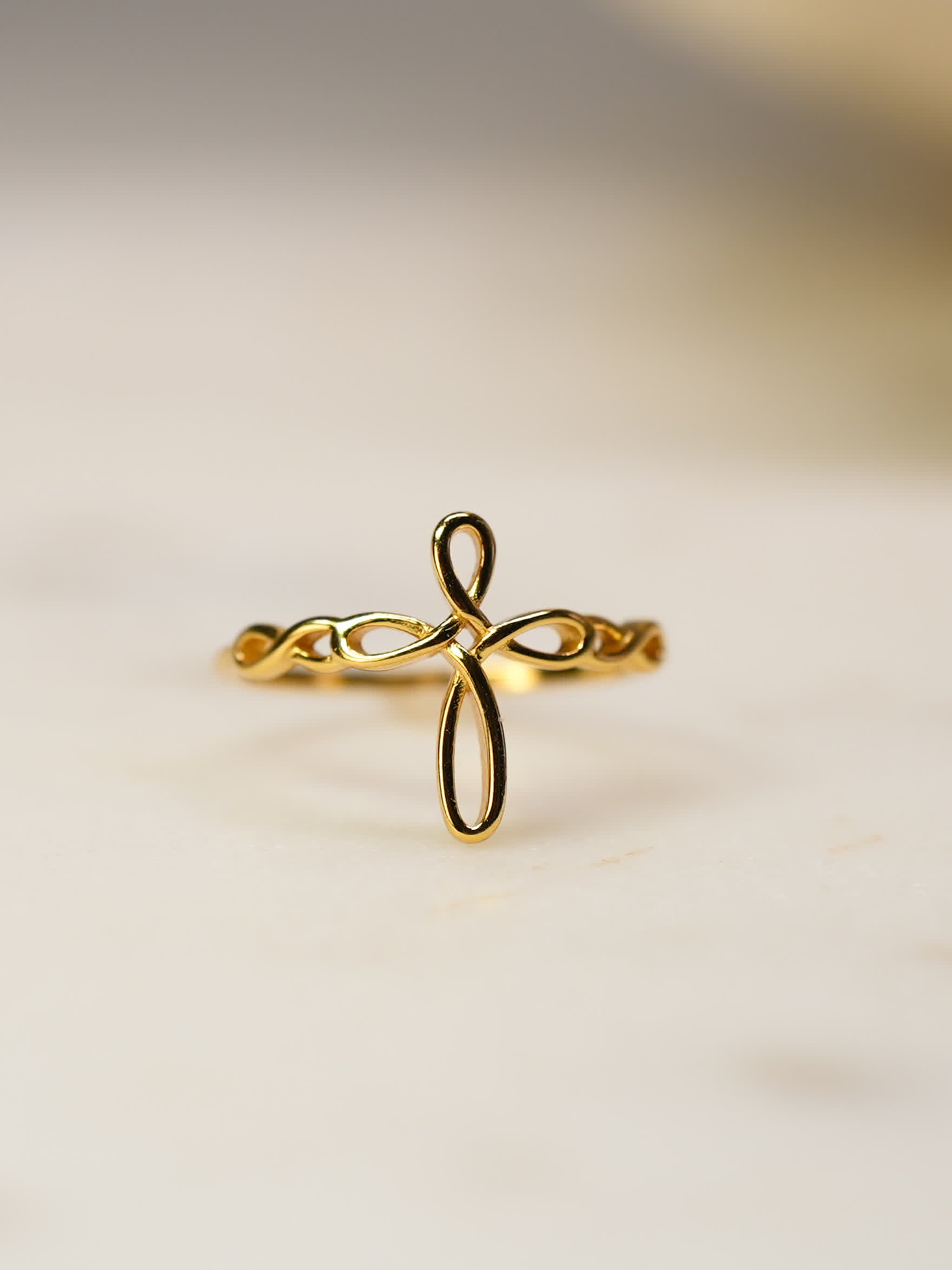 Close-up of gold ribbon faith ring resting on soft marble surface, highlighting flowing cross silhouette