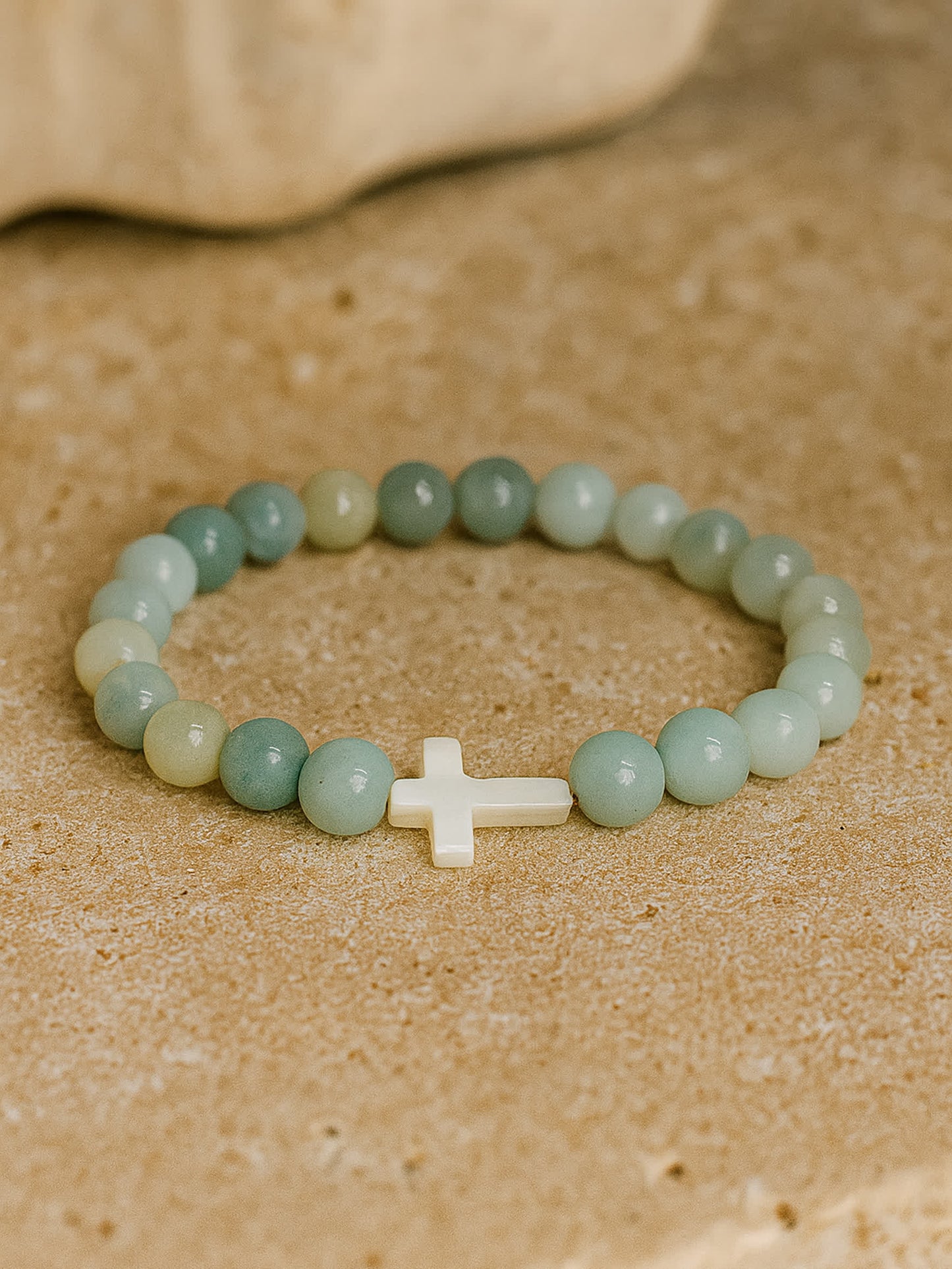 Flat lay photo of the Sea of Grace Cross Bracelet placed on a tan stone surface, evoking a natural, beach-like aesthetic with earthy warmth.