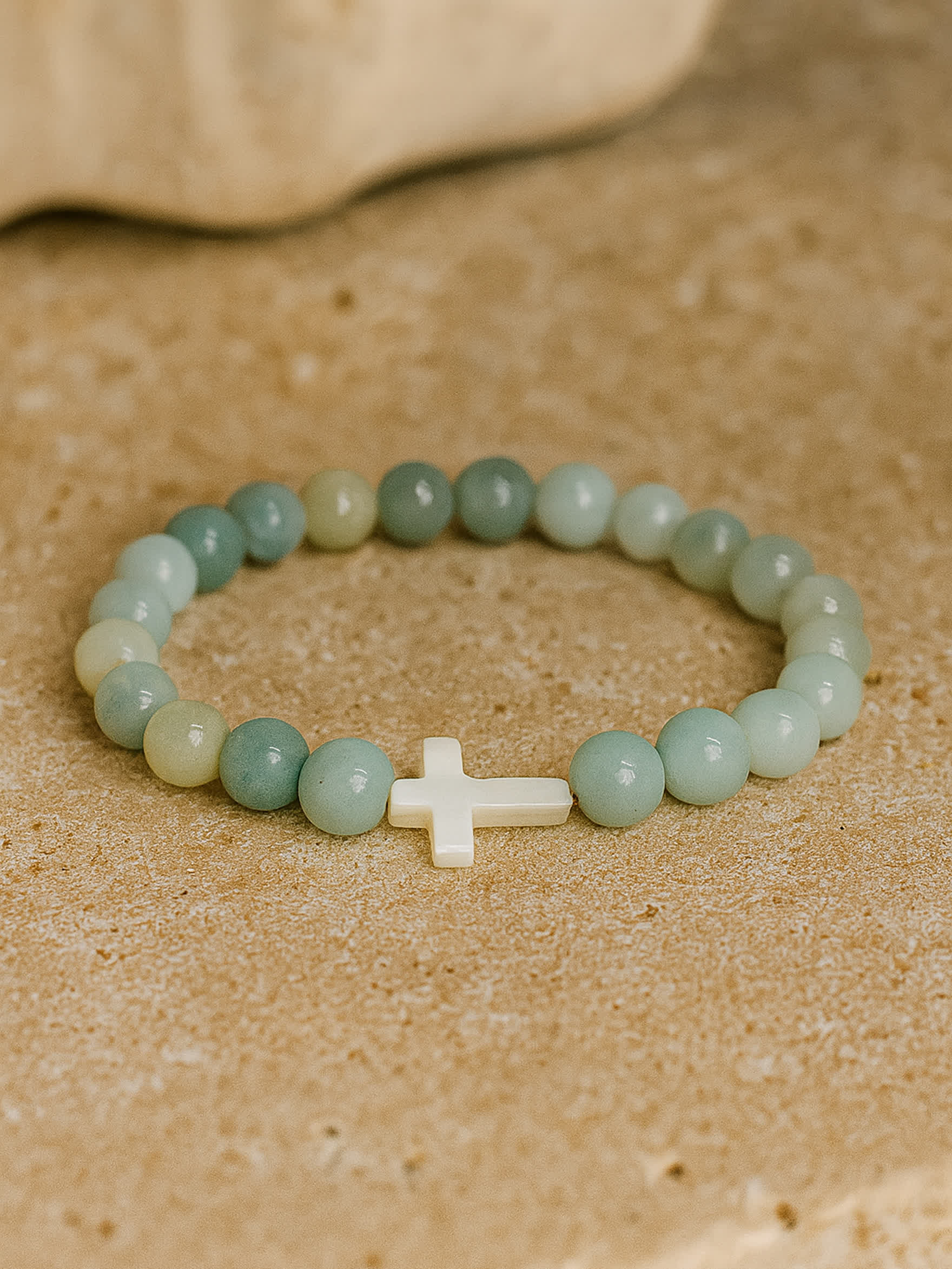 Flat lay photo of the Sea of Grace Cross Bracelet placed on a tan stone surface, evoking a natural, beach-like aesthetic with earthy warmth.