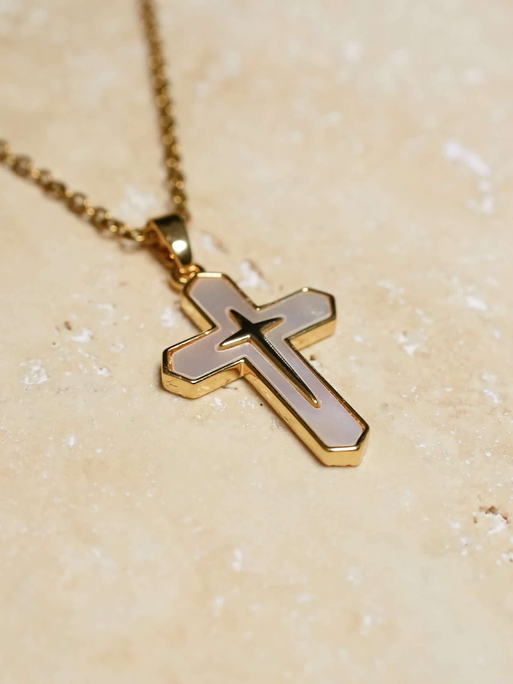 Gold and white cross necklace resting on a textured beige stone surface