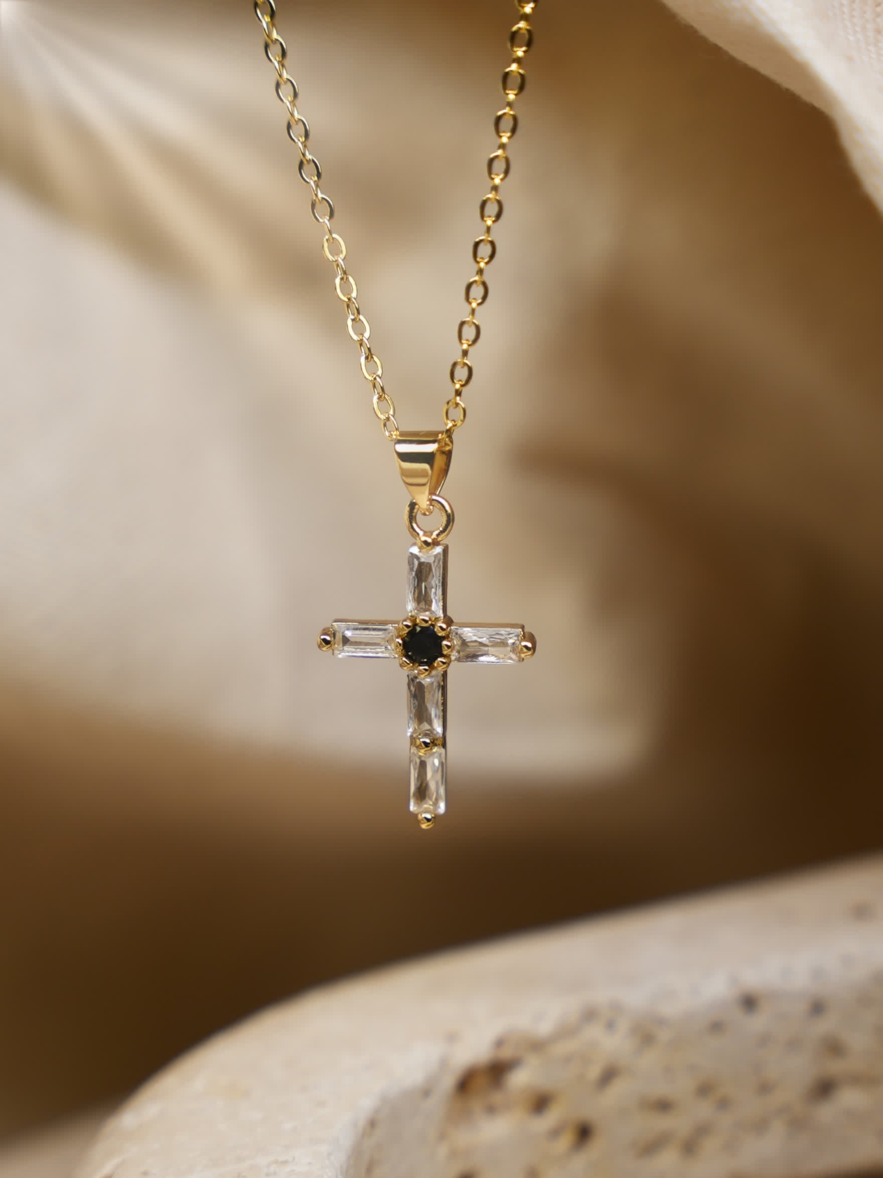 Gold cross necklace near soft fabric — A gold crystal cross necklace hanging upright in front of light beige fabric folds, giving a warm, elegant backdrop.