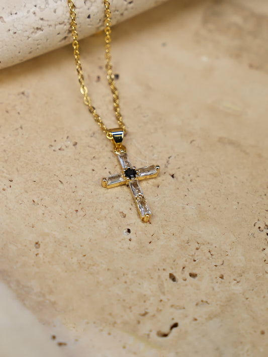 Gold cross necklace on travertine — A close-up of a gold baguette crystal cross necklace with black center stone, laid on a textured stone surface in natural light.