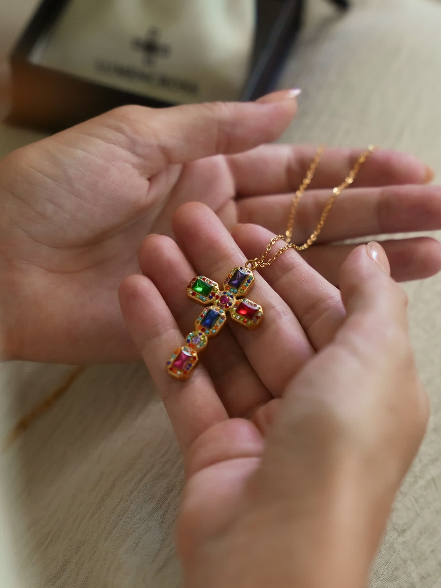 Two hands gently holding the Baroque multicolor gemstone cross necklace, emphasizing the pendant's vibrant jewel tones and sculptural gold frame.