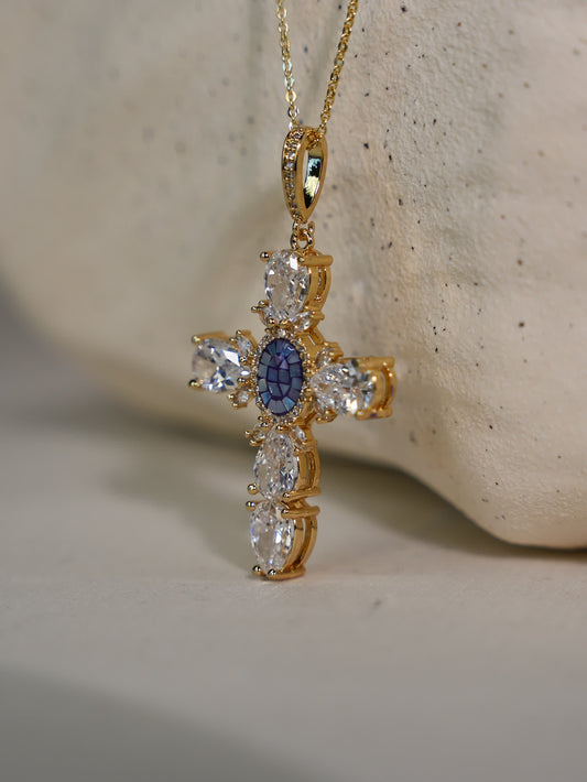 Hands gently holding the vintage-style gold cross necklace with sapphire-blue center stone and shimmering white accents