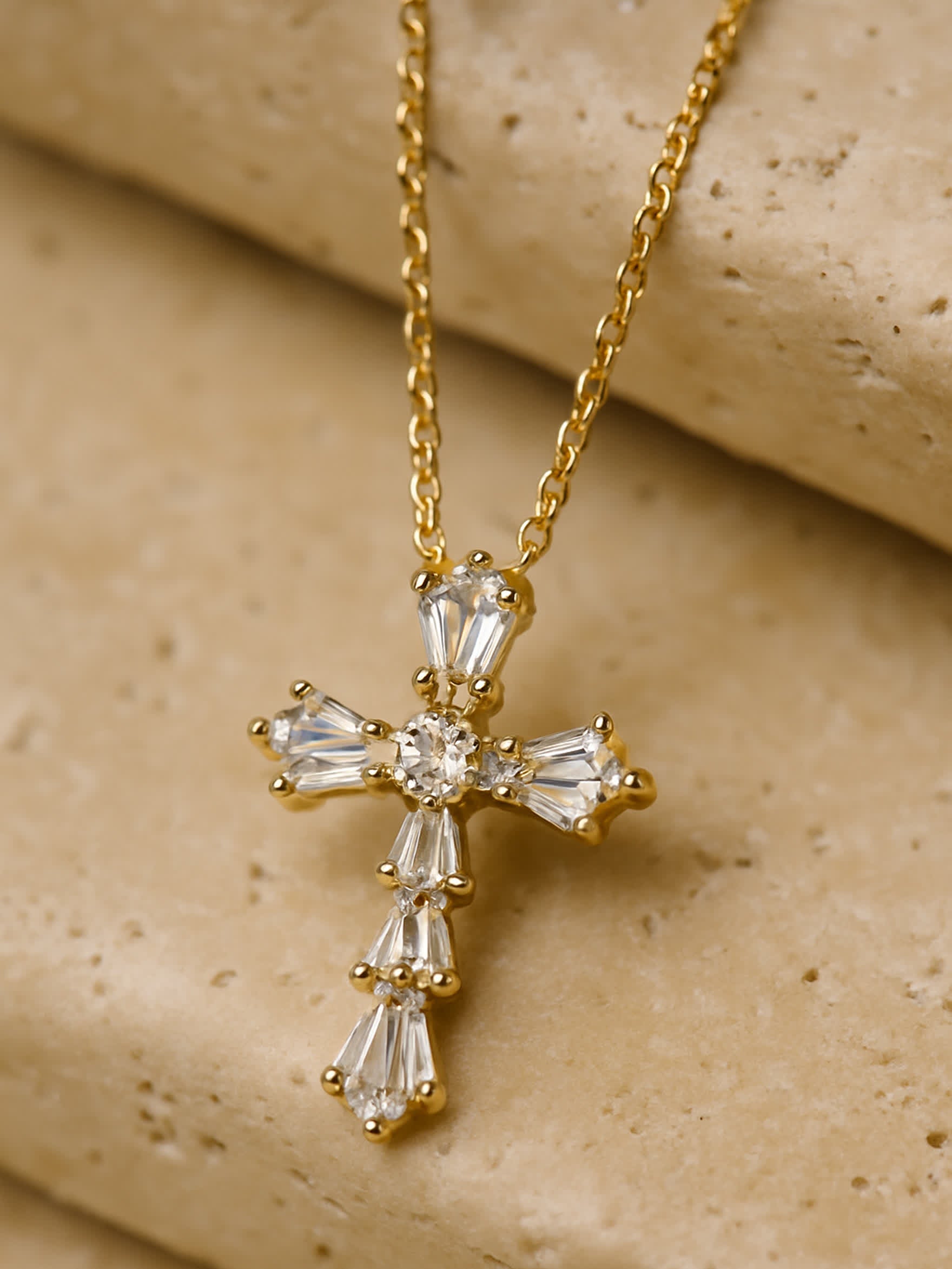 Close-up of Divine Bloom gold crystal cross necklace on travertine stone background, showcasing baguette and pear-cut zircon details