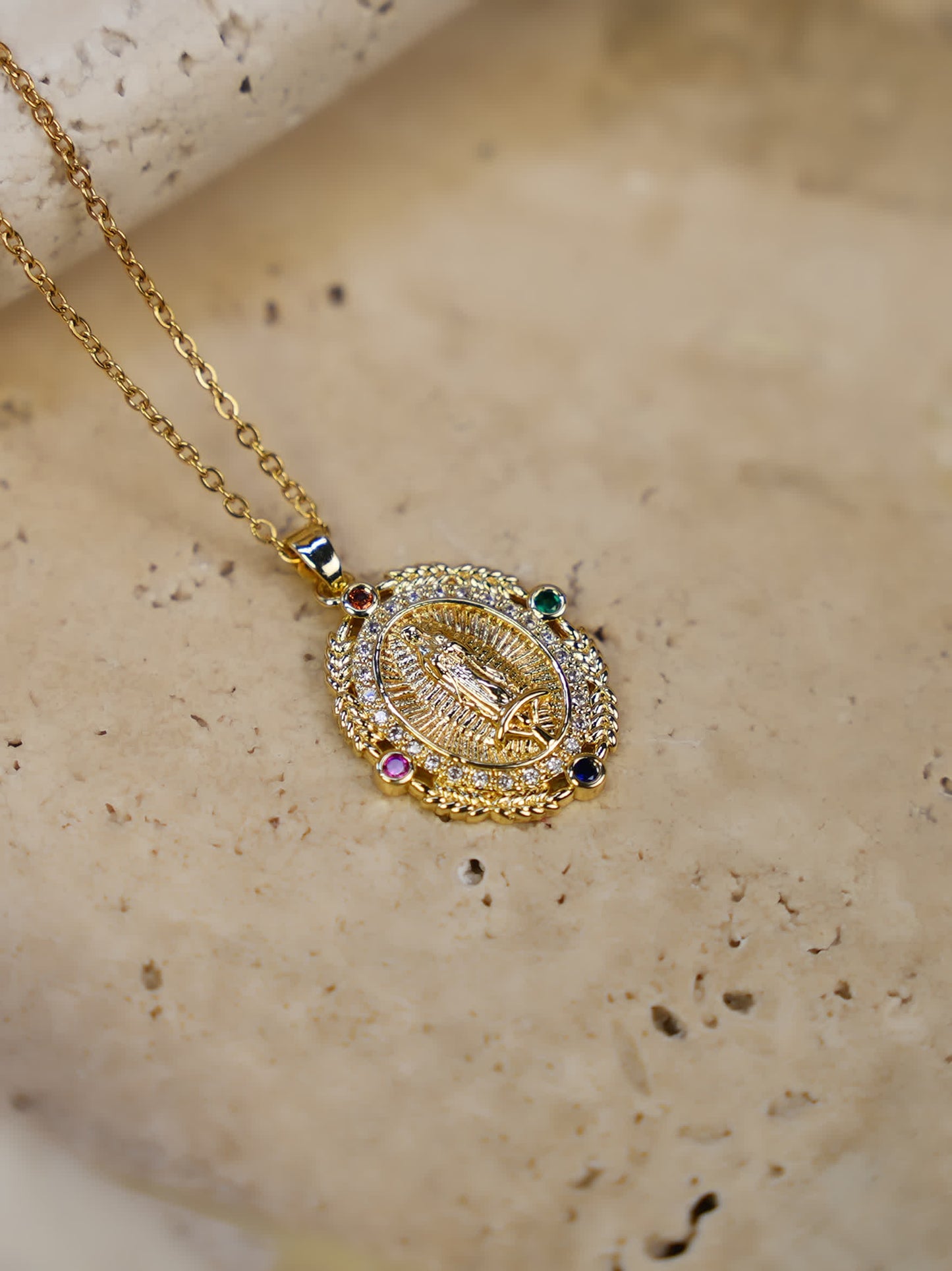 Virgin Mary medallion necklace resting on textured travertine, showcasing colorful stones and gold detailing