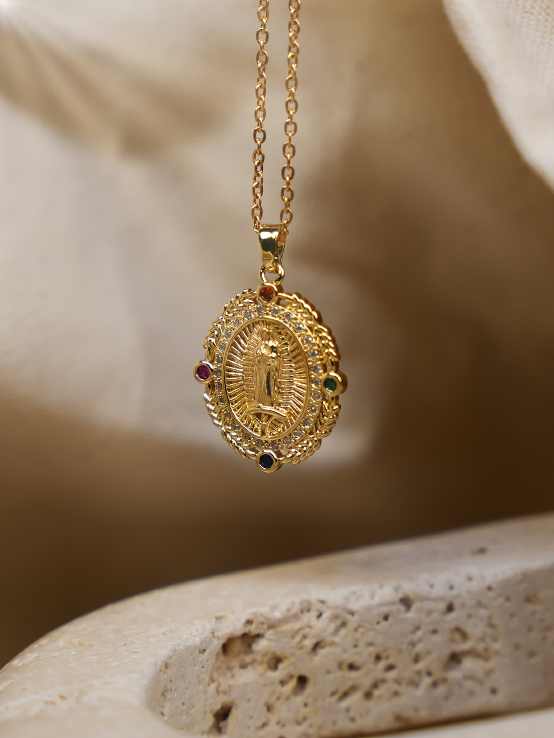 Gold Virgin Mary pendant necklace hanging over a beige stone surface with soft natural lighting