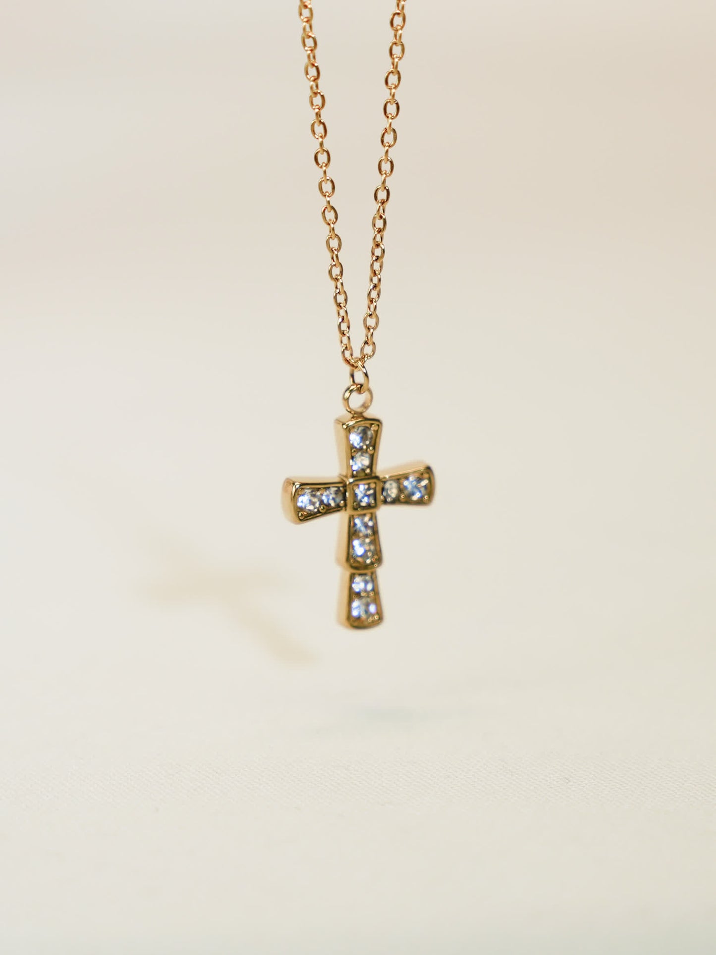 Front-facing product photo of a gold crystal cross necklace hanging on a delicate chain, captured against a soft beige background