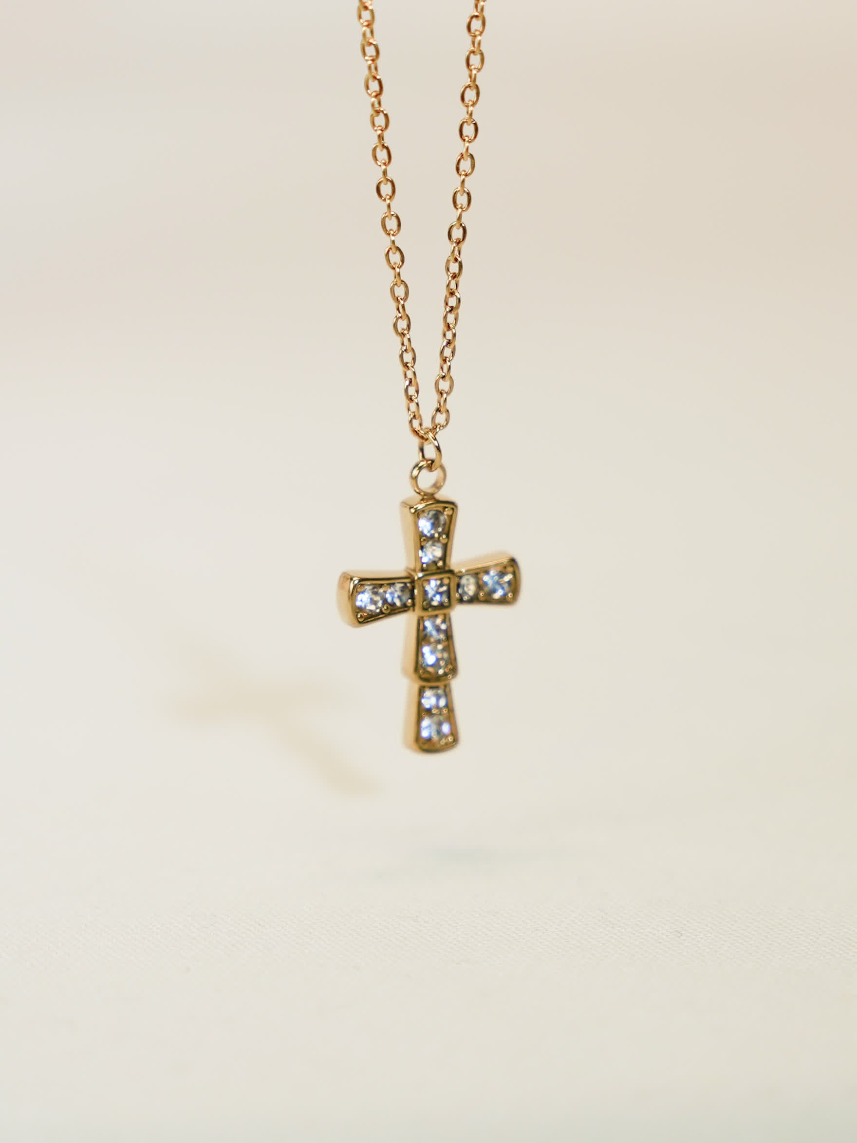 Front-facing product photo of a gold crystal cross necklace hanging on a delicate chain, captured against a soft beige background