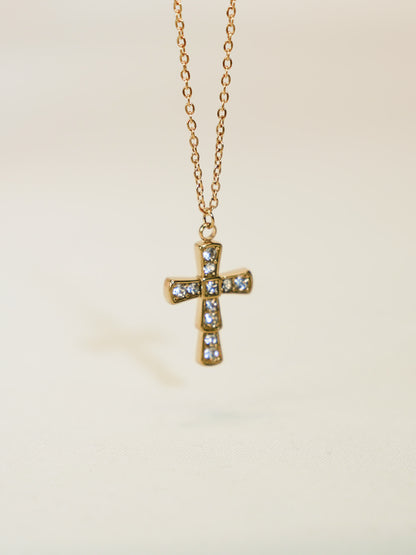 Front-facing product photo of a gold crystal cross necklace hanging on a delicate chain, captured against a soft beige background