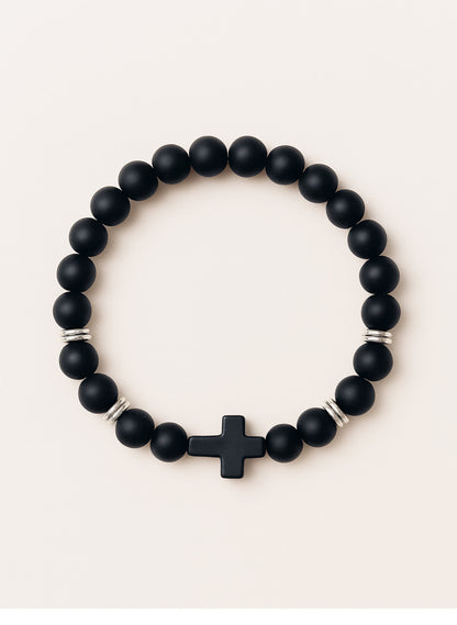 Studio product photo of the Covenant Shield Men's Cross Bracelet with matte black gemstone beads and a centered black cross, displayed against a soft cream background.