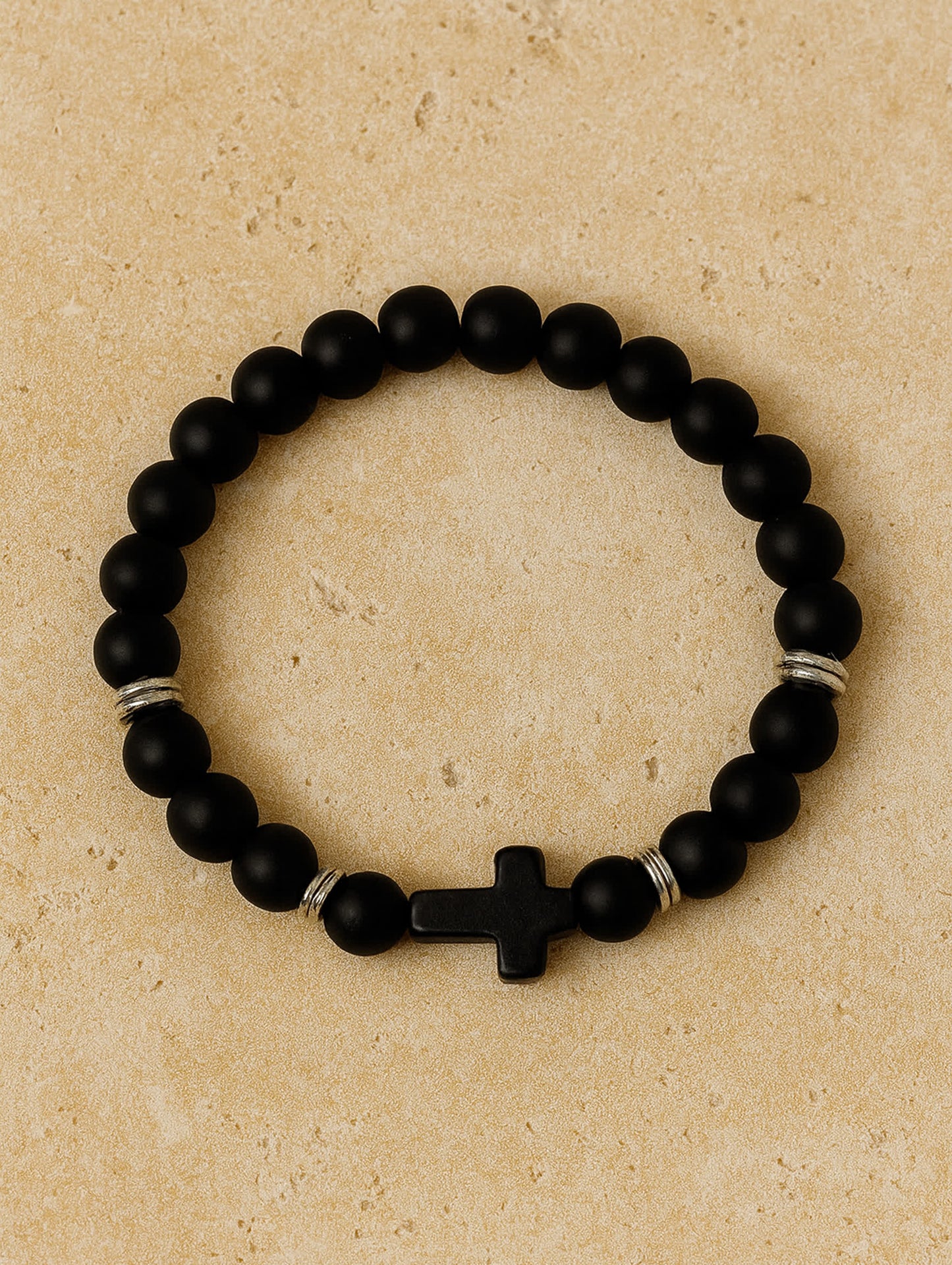 Overhead product image of the Covenant Shield Men's Cross Bracelet placed on a tan travertine surface, highlighting its sideways black cross and minimalist design.