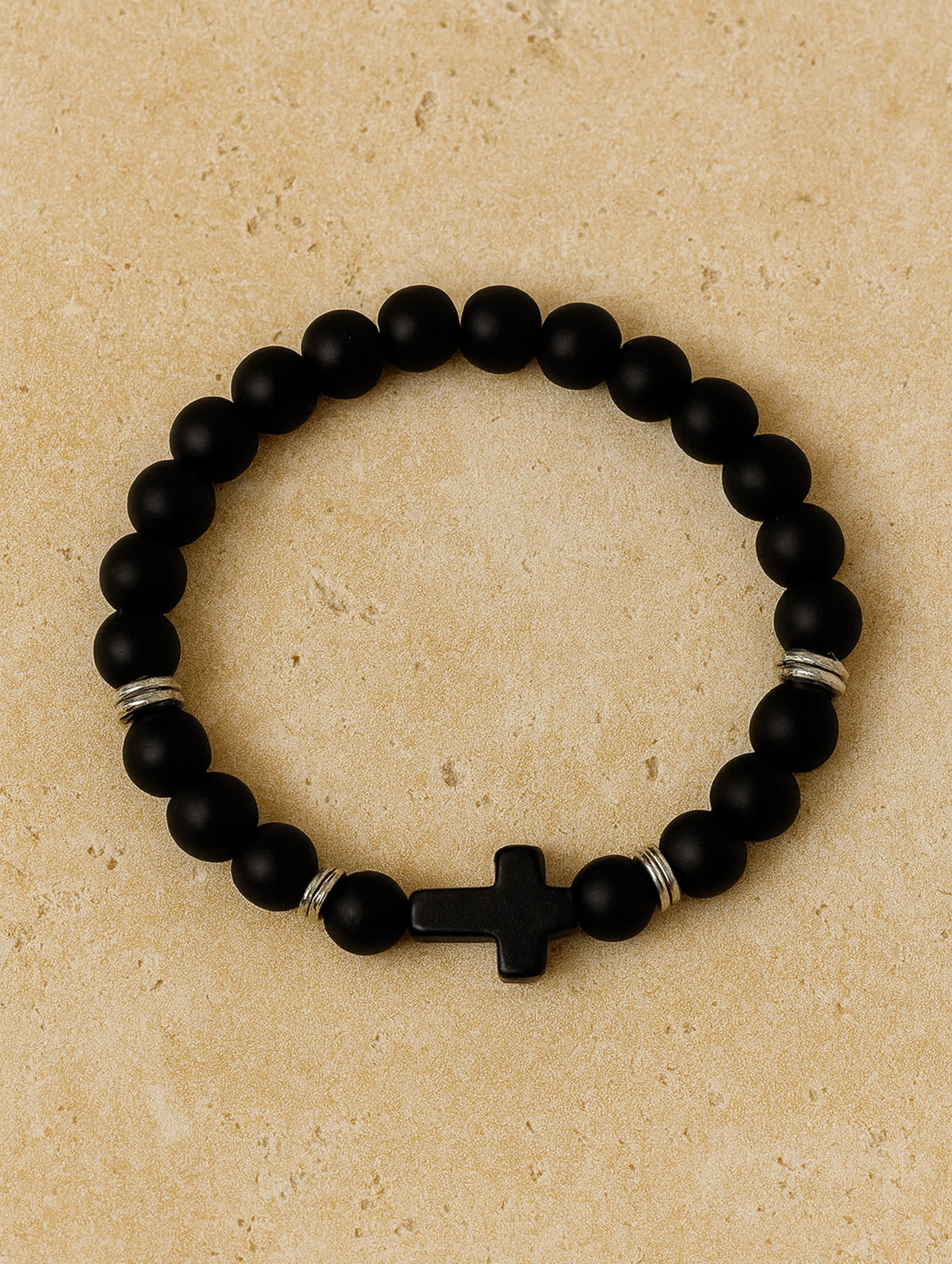 Overhead product image of the Covenant Shield Men's Cross Bracelet placed on a tan travertine surface, highlighting its sideways black cross and minimalist design.