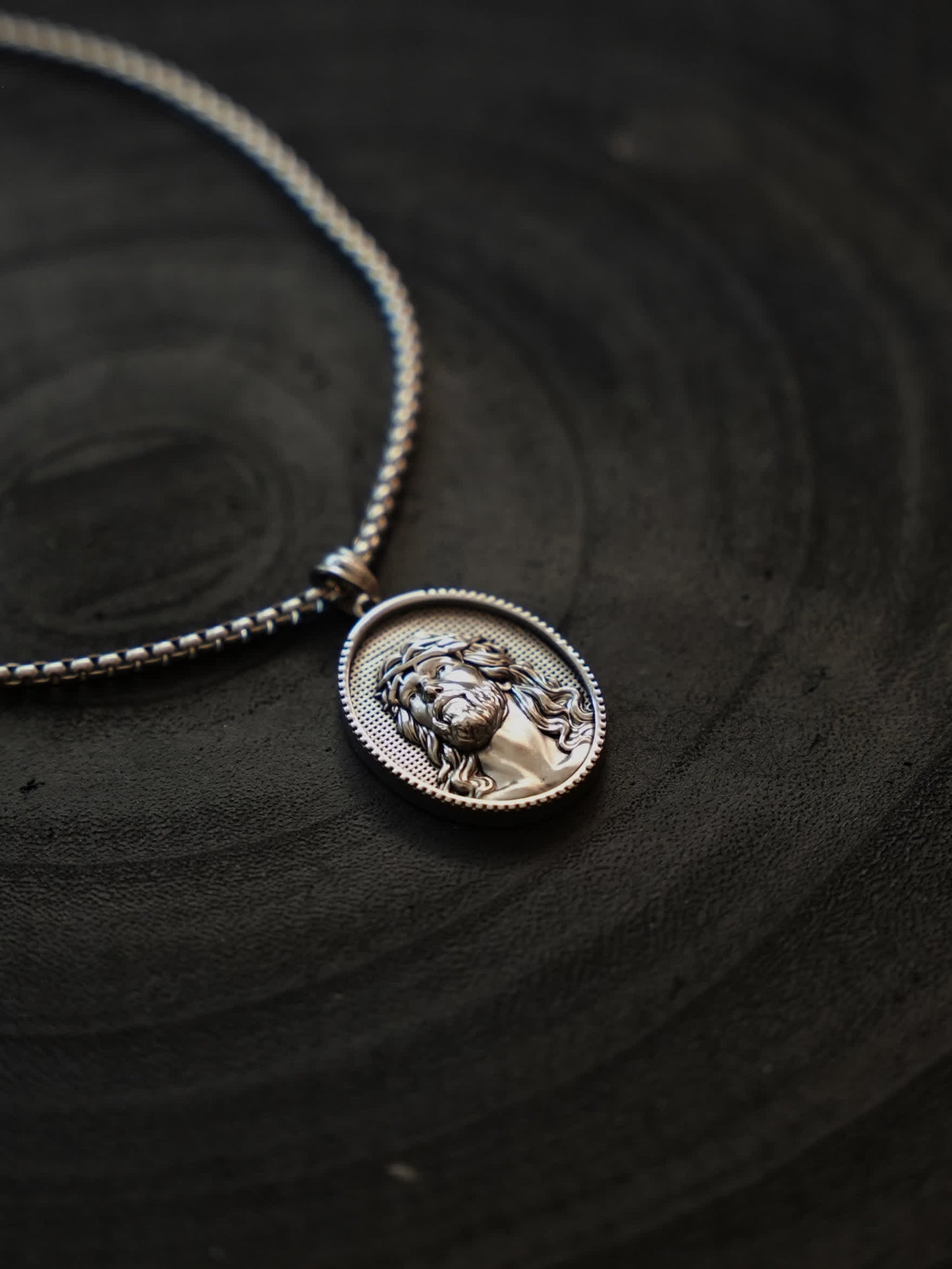 Christian medallion necklace laid flat on dark slate, emphasizing the contrast between the bold chain and engraved Jesus figure.