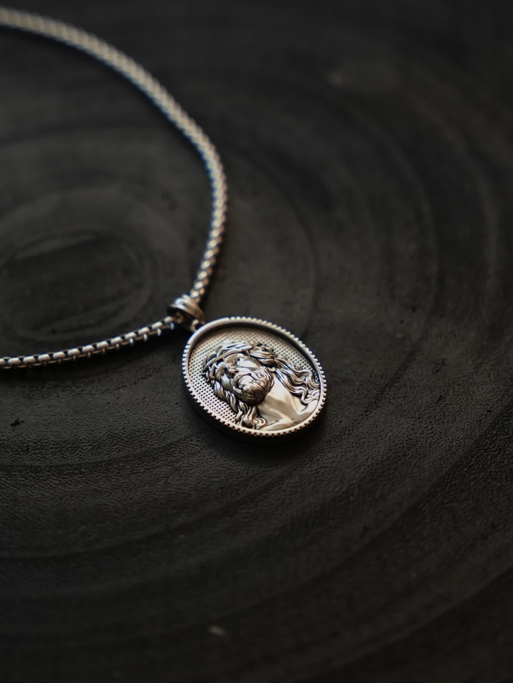 Christian medallion necklace laid flat on dark slate, emphasizing the contrast between the bold chain and engraved Jesus figure.