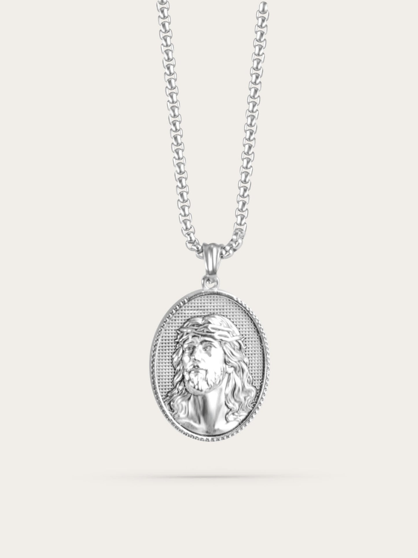 Front-facing studio photo of an oval Jesus face necklace with a polished stainless steel finish and crown of thorns design, displayed against a light beige background.
