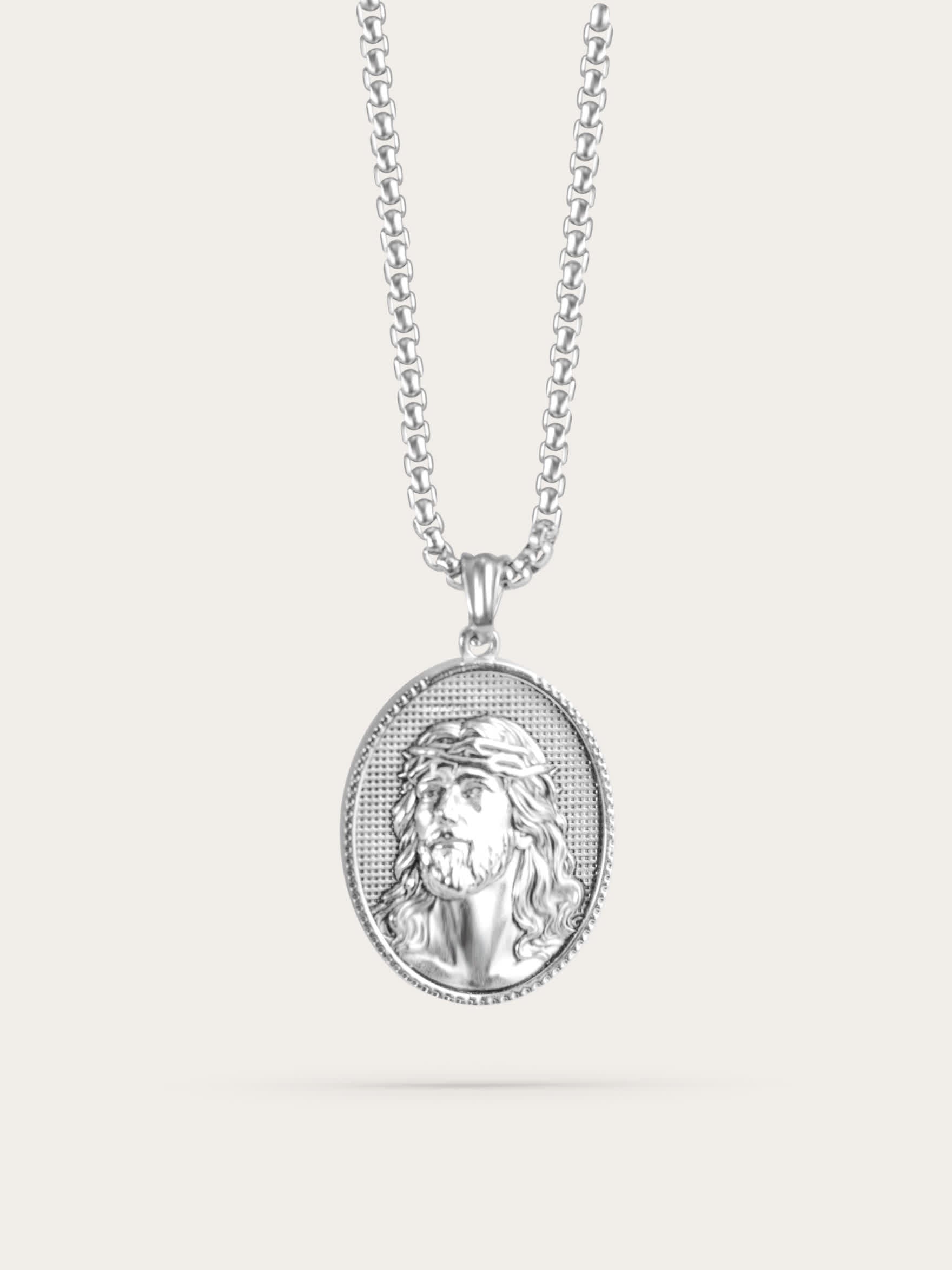 Front-facing studio photo of an oval Jesus face necklace with a polished stainless steel finish and crown of thorns design, displayed against a light beige background.