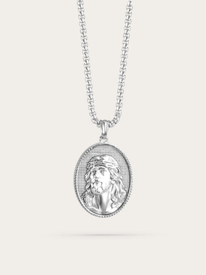 Front-facing studio photo of an oval Jesus face necklace with a polished stainless steel finish and crown of thorns design, displayed against a light beige background.