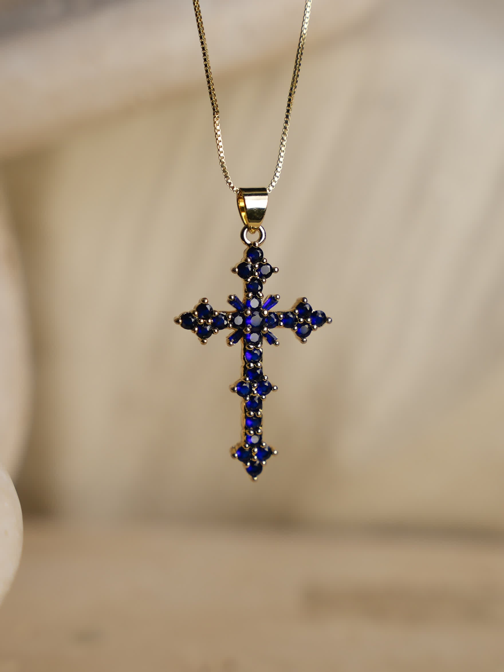 Close-up of the blue cross necklace resting on neutral fabric background, showcasing deep blue zircon stones and gold chain