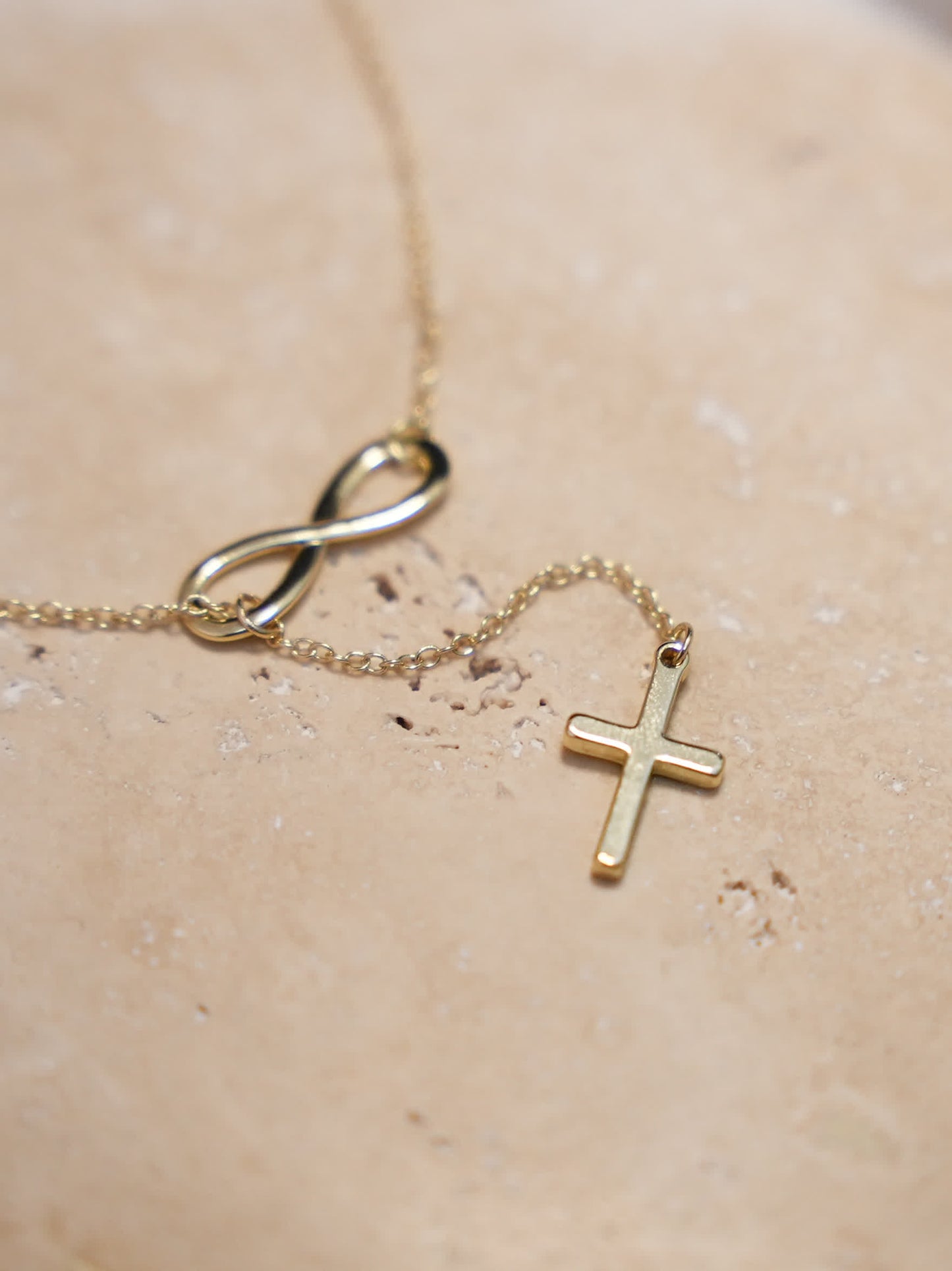 Overhead product photo of the necklace on a beige stone slab, with both the infinity loop and cross clearly visible