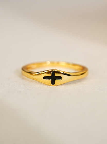 Sculpted signet-style gold ring with recessed black cross, displayed on soft ivory fabric