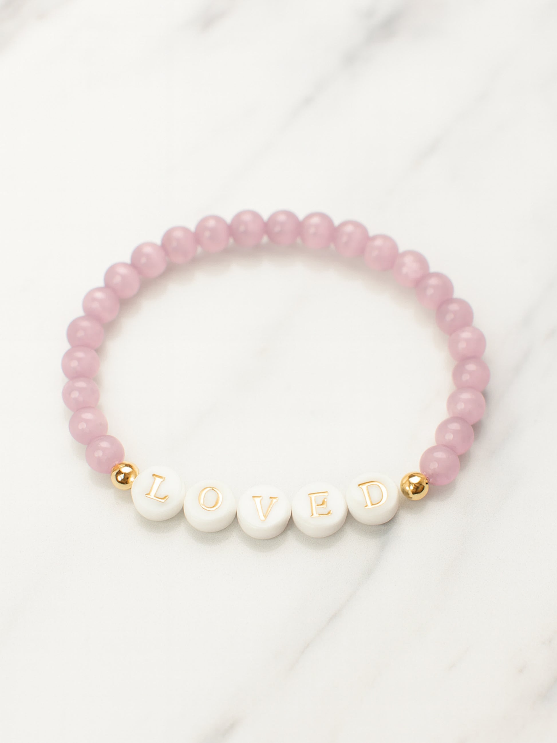Flat lay product photo of the Heaven’s Whisper Faith Bracelet arranged on a white marble surface, showing the full bracelet design with soft pink beads and gold-detailed letter accents.