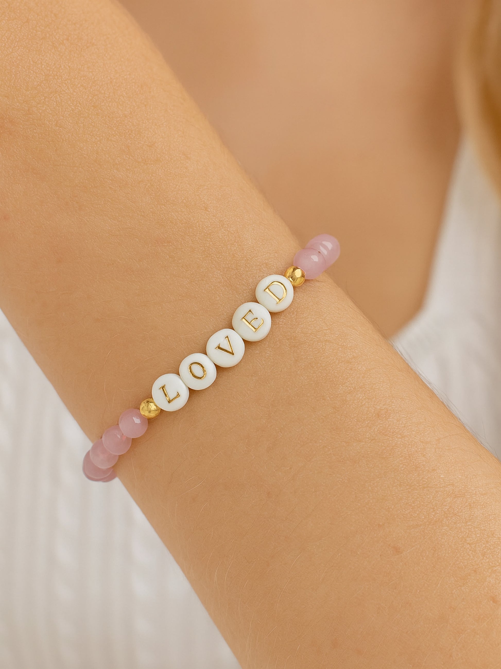 Lifestyle image of the Heaven’s Whisper Faith Bracelet worn on a woman's wrist, featuring pink Cat’s Eye beads and white letter beads spelling "LOVED" with gold accents.