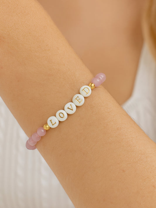 Lifestyle image of the Heaven’s Whisper Faith Bracelet worn on a woman's wrist, featuring pink Cat’s Eye beads and white letter beads spelling "LOVED" with gold accents.