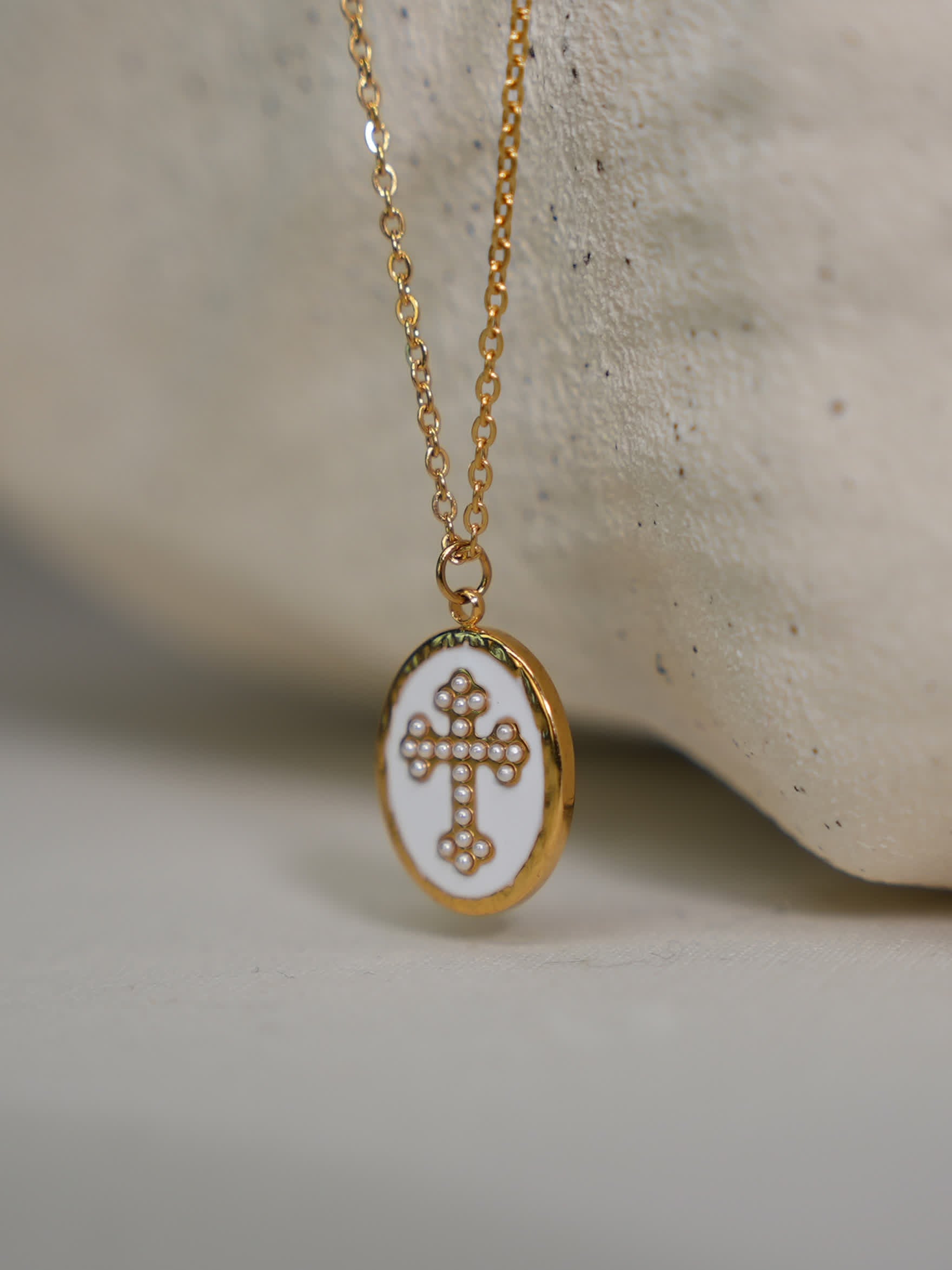 Pendant leaning on light stone texture
Close-up of gold and white enamel cross necklace draped over curved stone