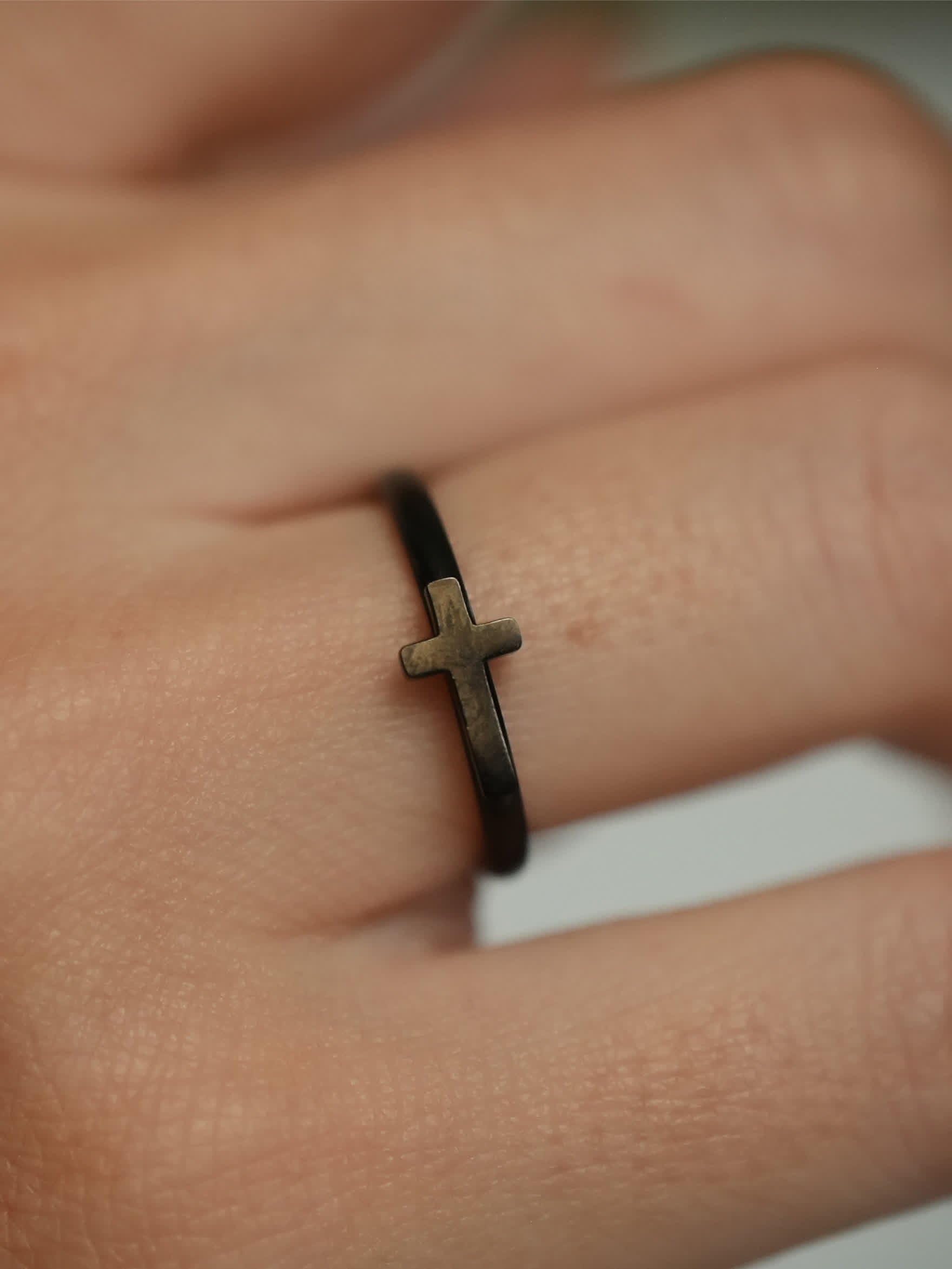 Macro side view of the woman’s cross ring, highlighting the antique bronze-tone cross detail against the matte black band on the finger.

