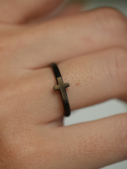 Macro side view of the woman’s cross ring, highlighting the antique bronze-tone cross detail against the matte black band on the finger.

