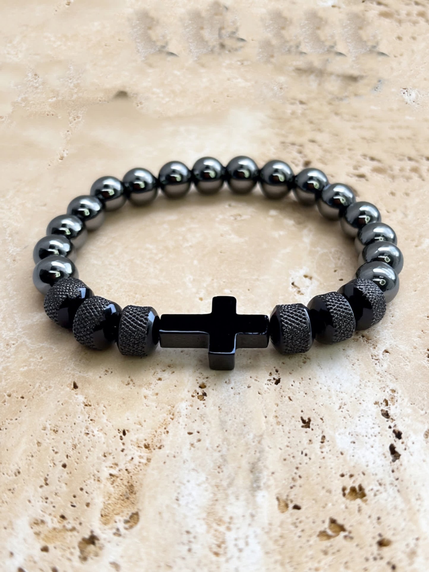 Overhead product shot of the Iron Grace Beaded Cross Bracelet on a natural travertine surface, showcasing the polished black cross and reflective bead finish.