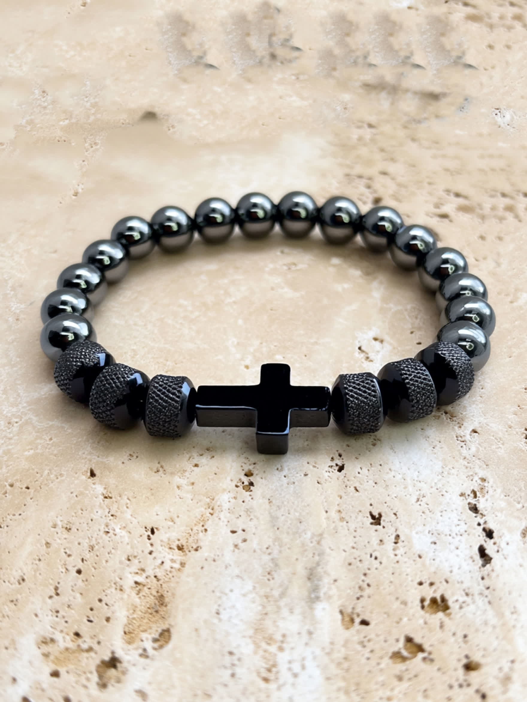 Overhead product shot of the Iron Grace Beaded Cross Bracelet on a natural travertine surface, showcasing the polished black cross and reflective bead finish.